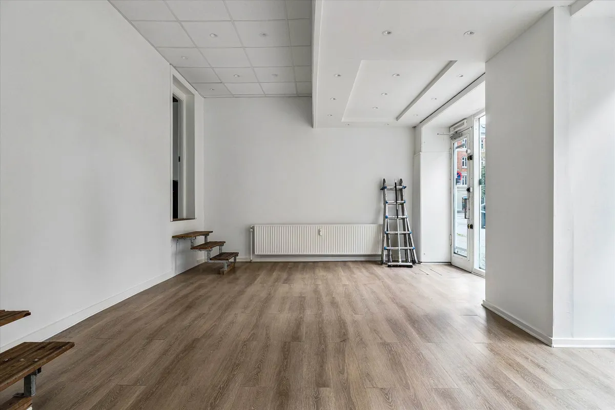 Et tomt showroom med højt til loftet og hårdt, trægulv i grå nuancer. Ståbord, trapper og en stige mod væggen