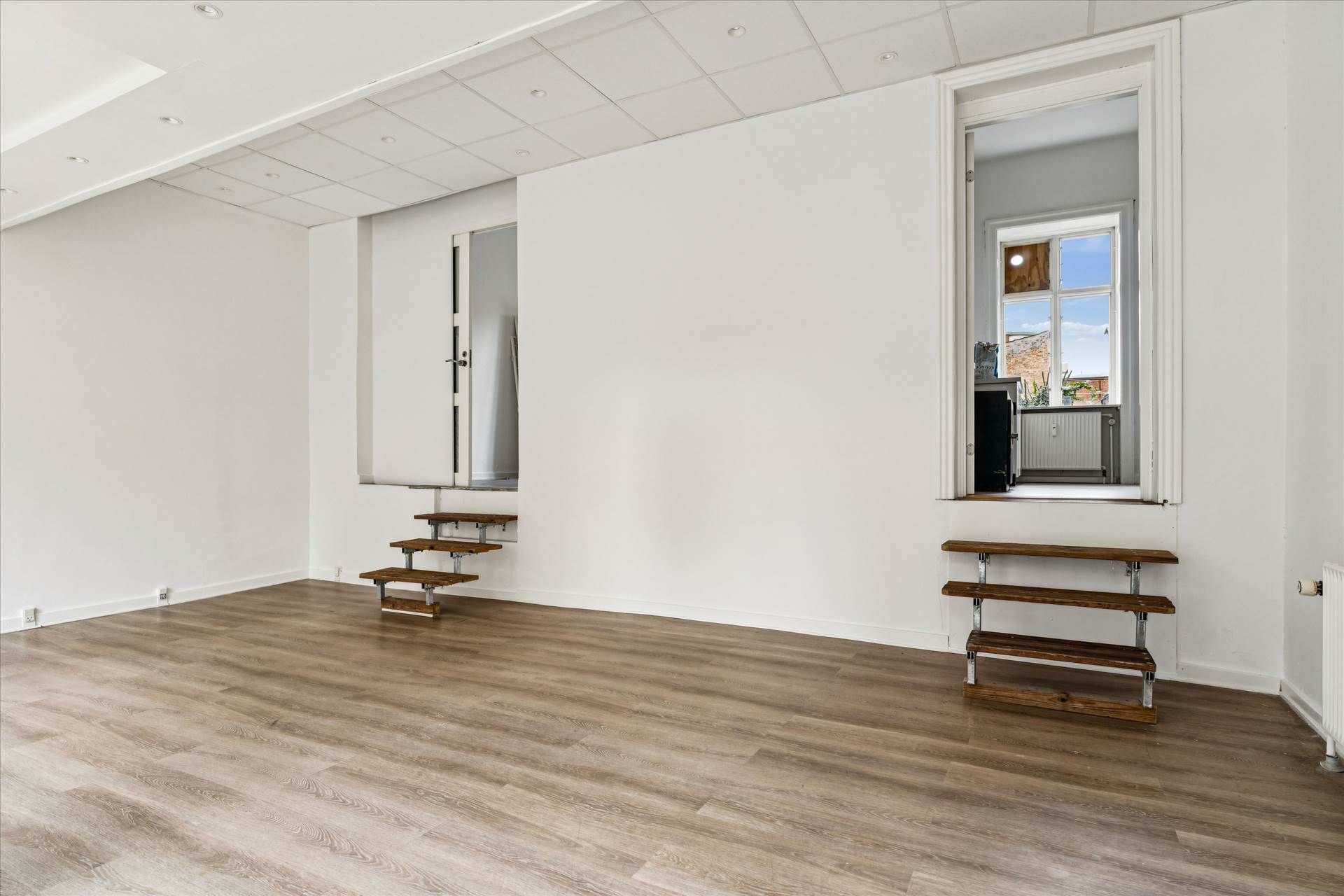 Et tomt showroom med højloft, parketgulv og to trætrapper, der fører op til et vindue. Et andet vindue og et dørkar og