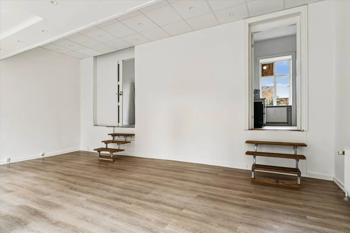 Et tomt showroom med højloft, parketgulv og to trætrapper, der fører op til et vindue. Et andet vindue og et dørkar og