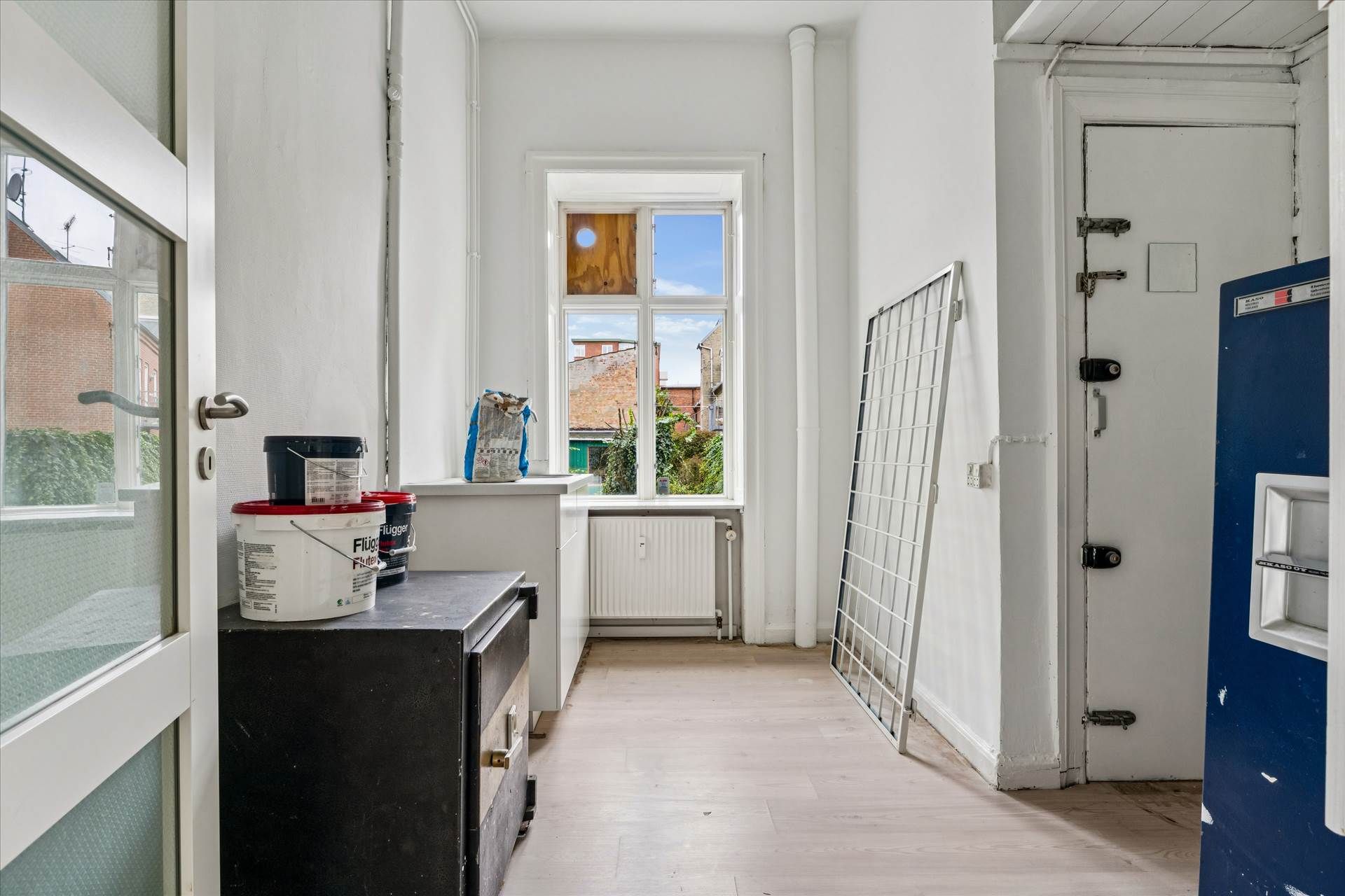 En tomt indre rum med en hvid væg og et vindue med et hul i midten, hvor der er en udsigt til bygninger og planter udenfor