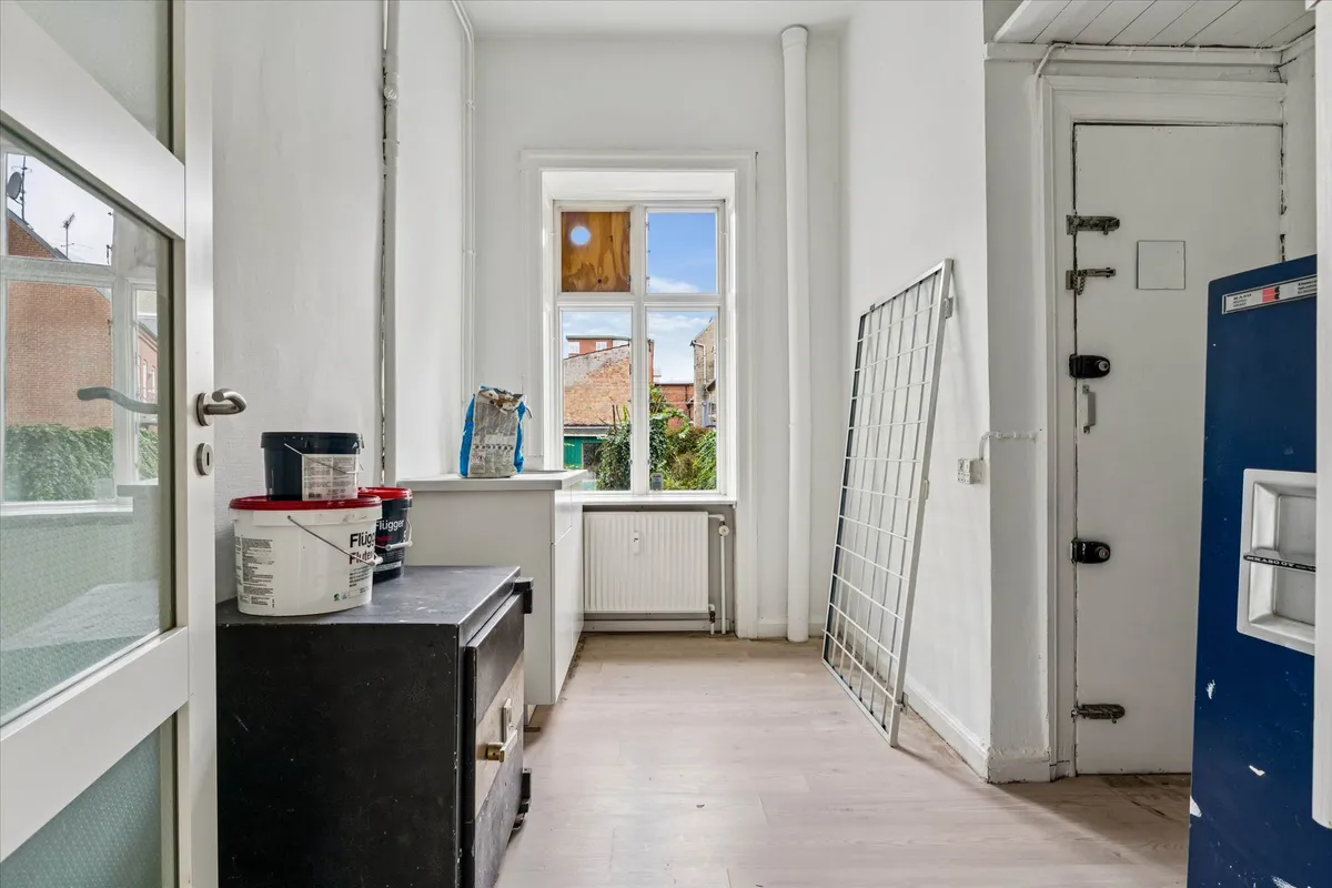 En tomt indre rum med en hvid væg og et vindue med et hul i midten, hvor der er en udsigt til bygninger og planter udenfor