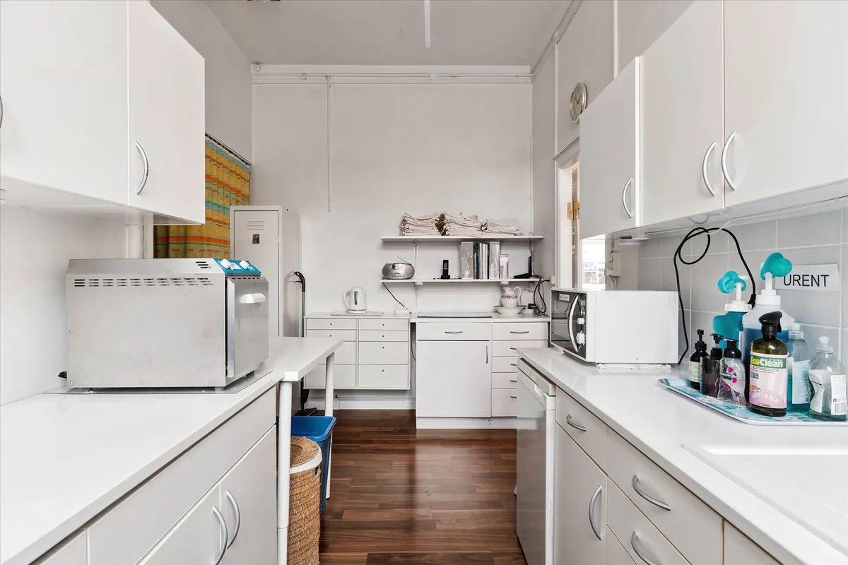 En ren og pæn køkkenrum med hvide køkkenskabe, to mikroovne, og et bord med rengøringsmidler.