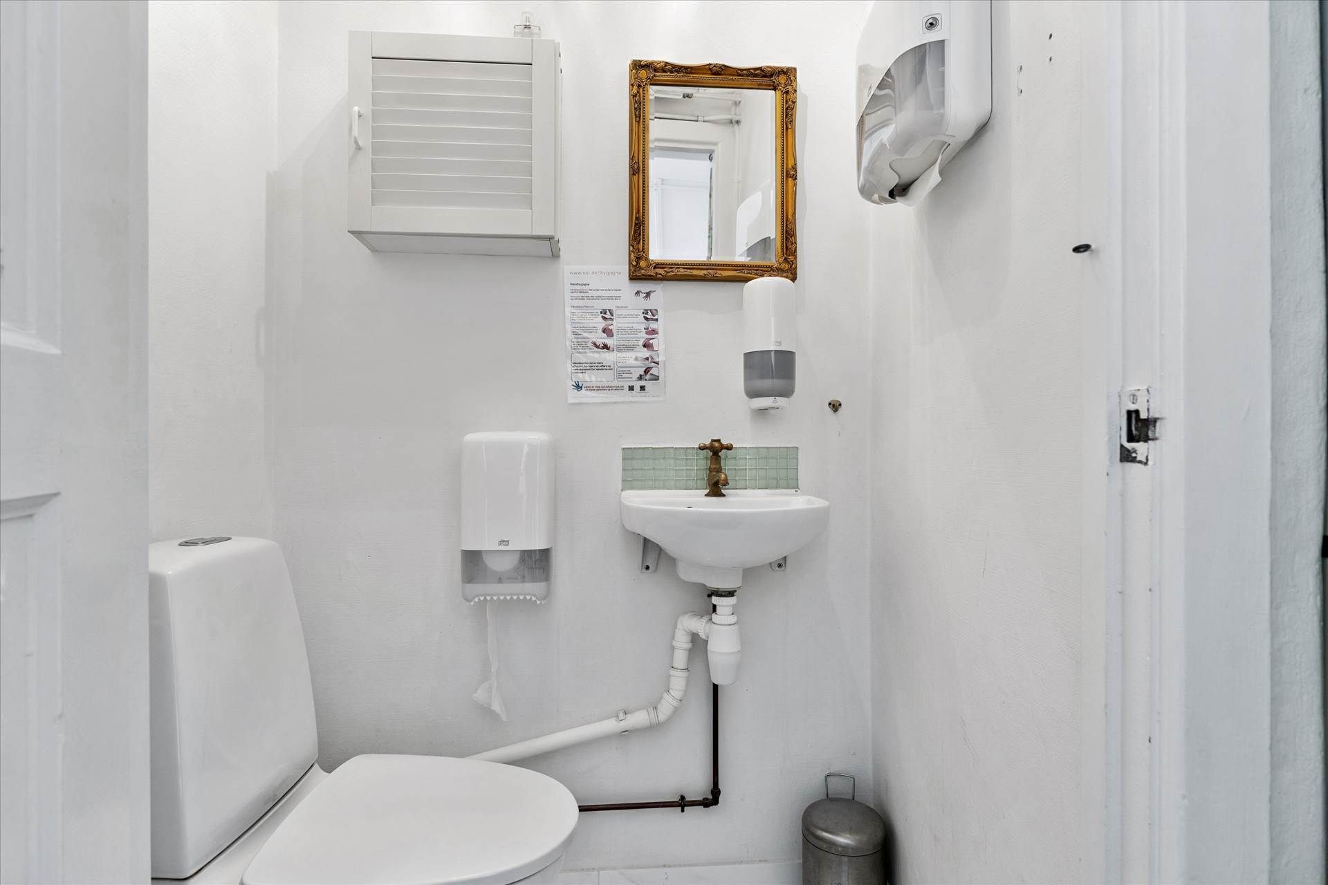 Et lille, hvidt badeværelse med toilet, håndvask og spejl. Væggen er hvid og der er et håndsæbe dispenser og en