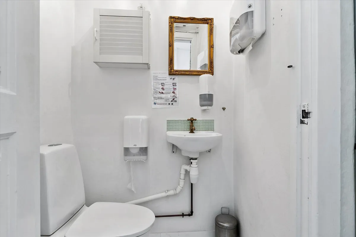 Et lille, hvidt badeværelse med toilet, håndvask og spejl. Væggen er hvid og der er et håndsæbe dispenser og en