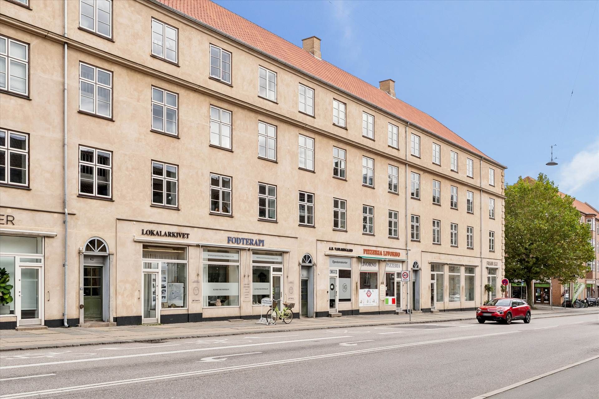 Bred gade med rødt nyttighedsvogn, to butikker, Lokalarkivet og Fodterapi, og cykelparkering i forgrunden