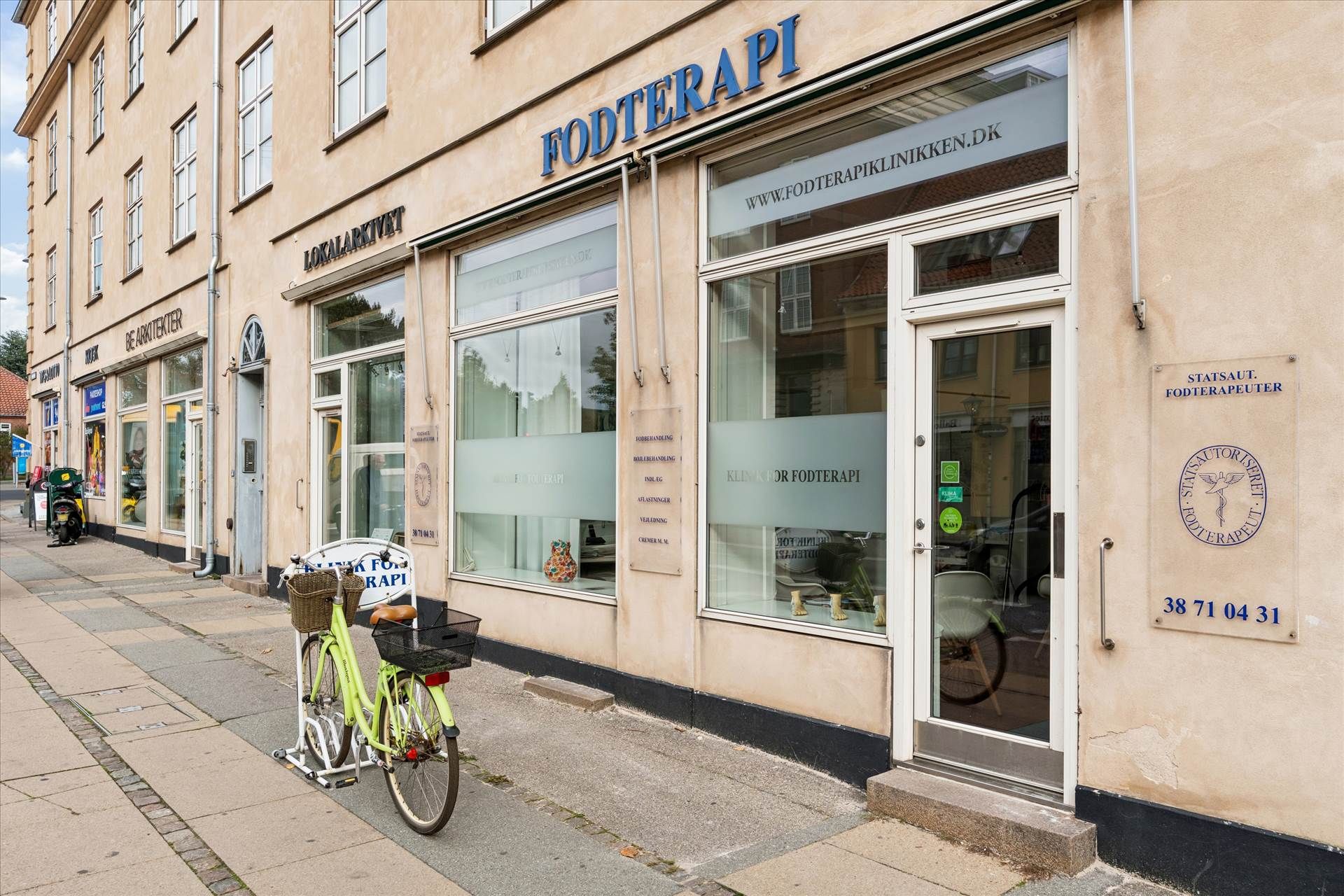 En grøn cykel parkeret foran en butik med navnet Fodterapi og teksten www.fodterapiklinikken.dk