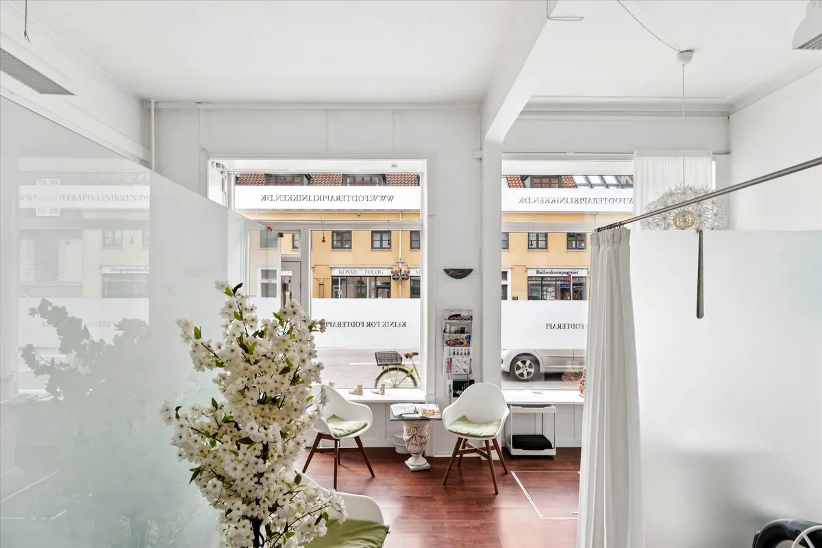 Interiør med hvide vægge, lyse lamper, to lænestolpe med grønne pude, blomster og åbent vindue med udsigt til gaden