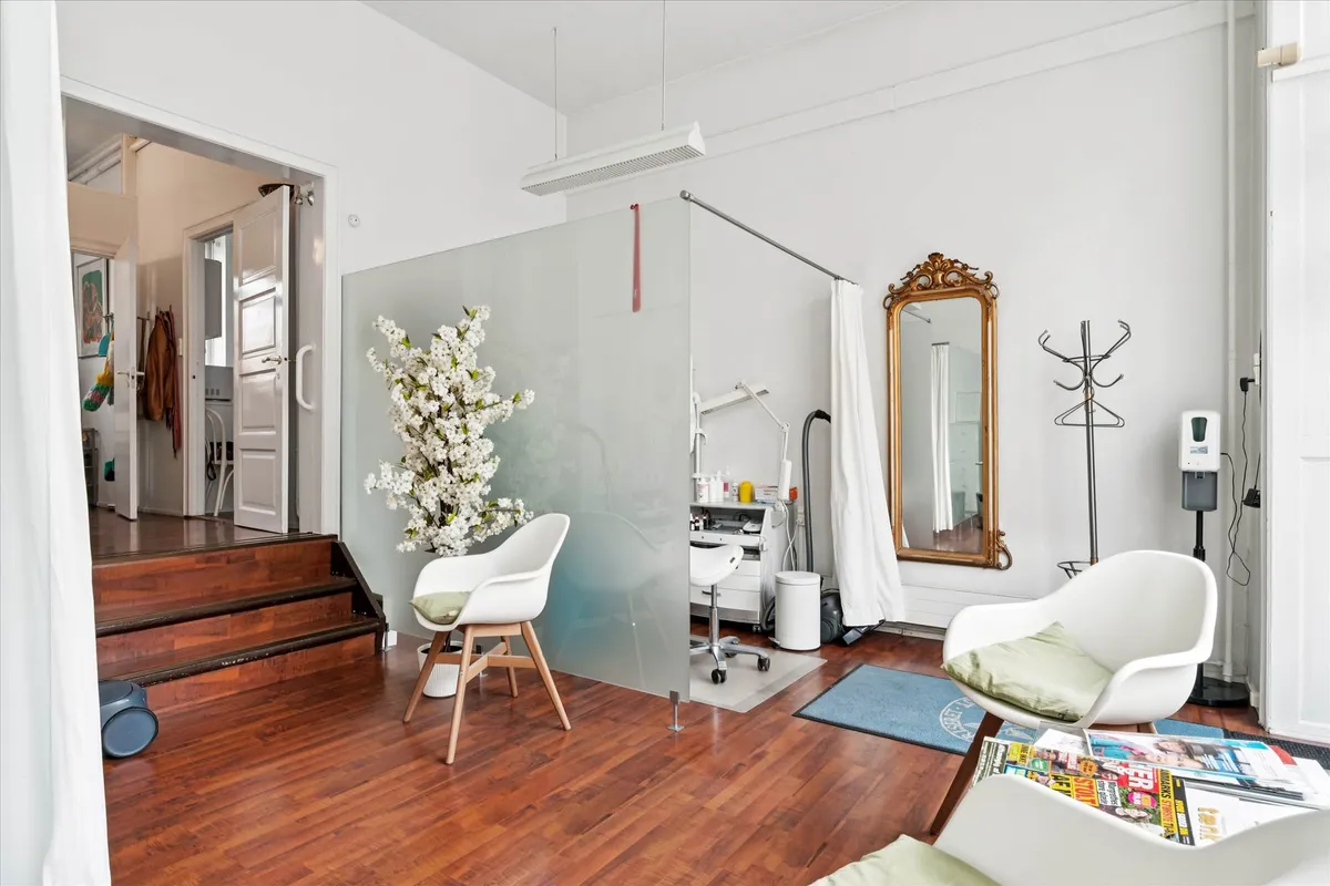 I et lyse værelse med hårde trægulve står en lysegrå lænestol ved siden af en blomsterpotte og en hvid sofa med grøn pude