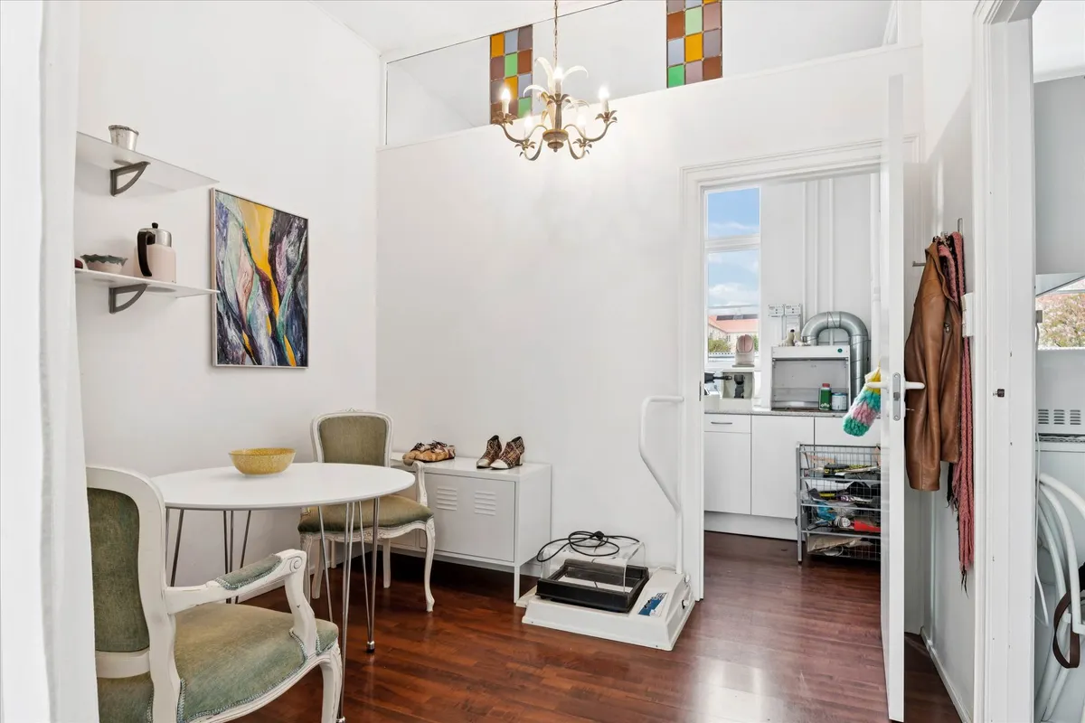 En hvid stue med et rundt bord og fire stole samt et skab med en skovl og en kedel på hylden