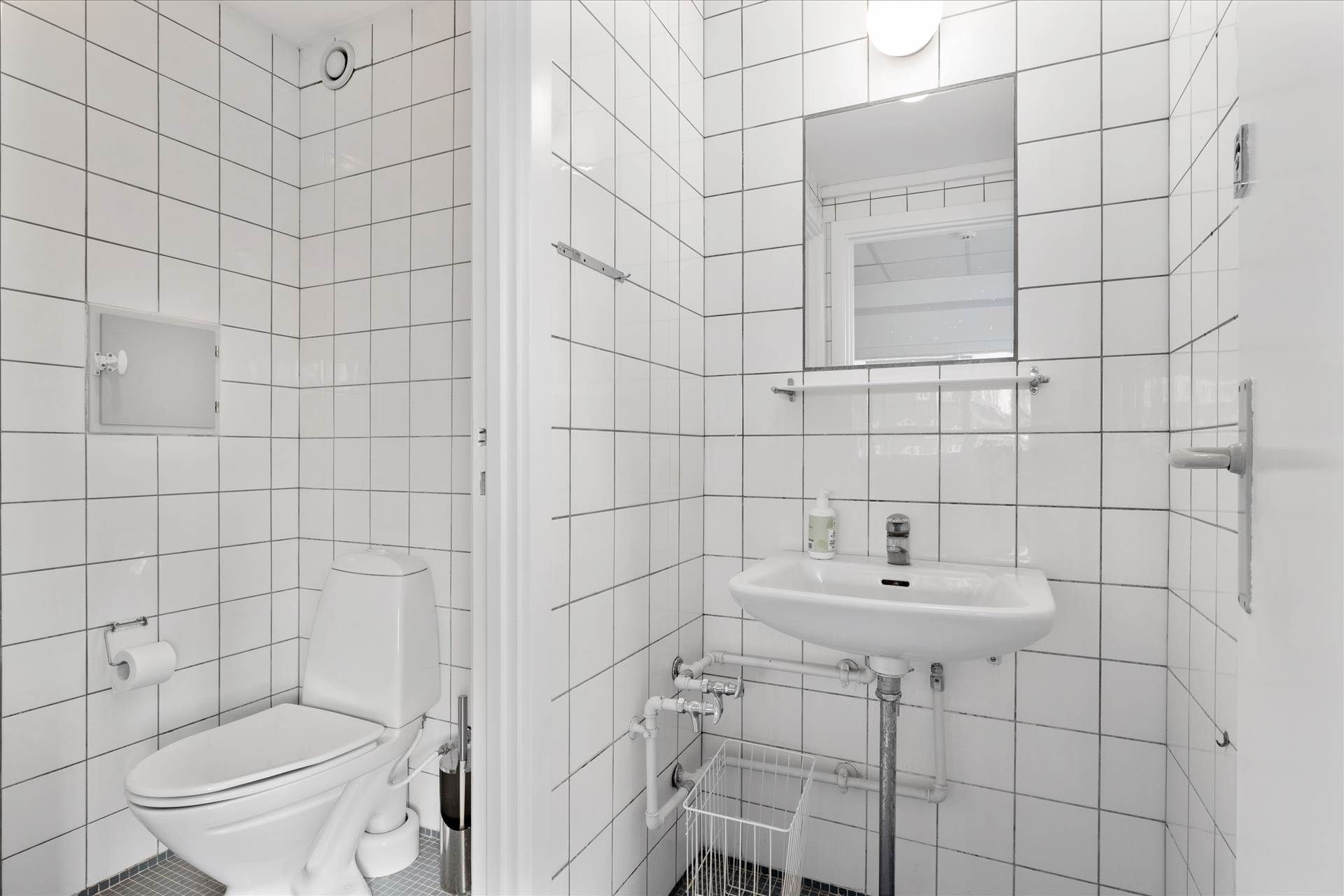 Et rent og velplejet badeværelse med hvidt toilet, vask og spejl samt gulv med mosaikfliser og hvide kakeløg