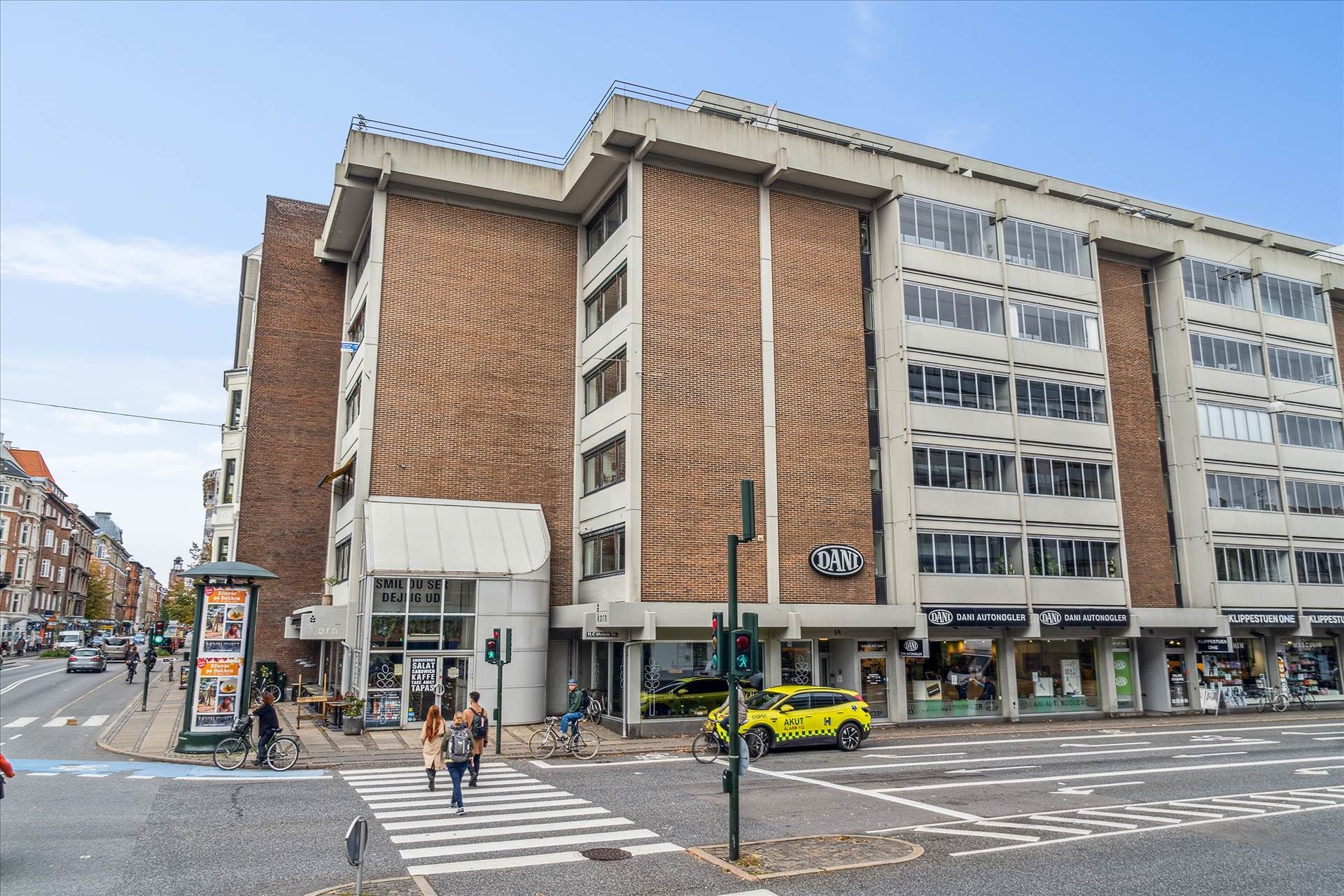 Kontorbygning med butikker på gadehjørnet, flere mennesker krydser på zebraovergangen og biler og cykler på vejen