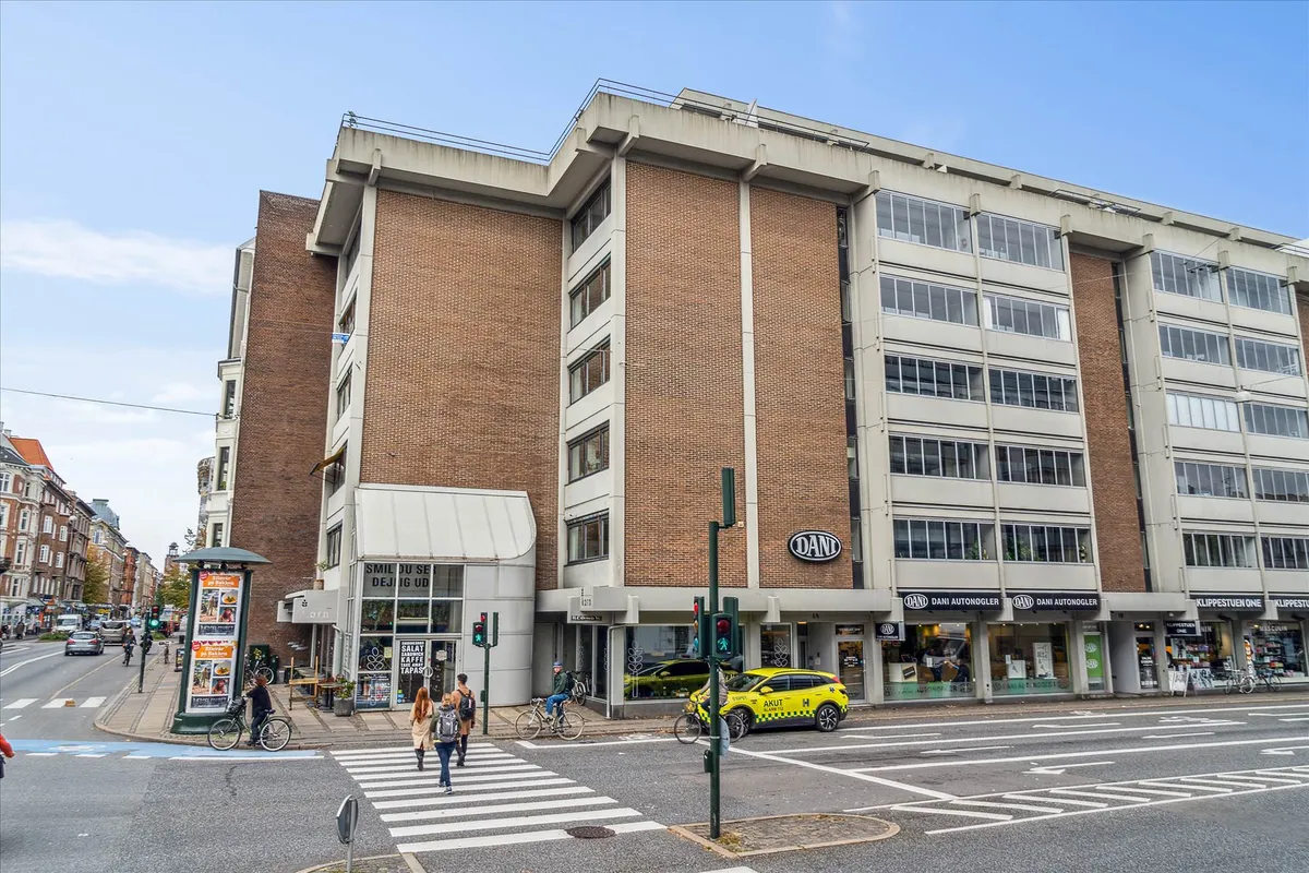 Kontorbygning med butikker på gadehjørnet, flere mennesker krydser på zebraovergangen og biler og cykler på vejen