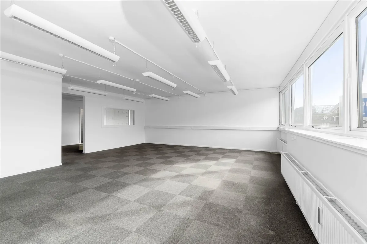 Et ubeboet og rent kontor med hvide vægge og gulv, hvidt loft og store vinduer, hvori der ses en bygning og et skilt