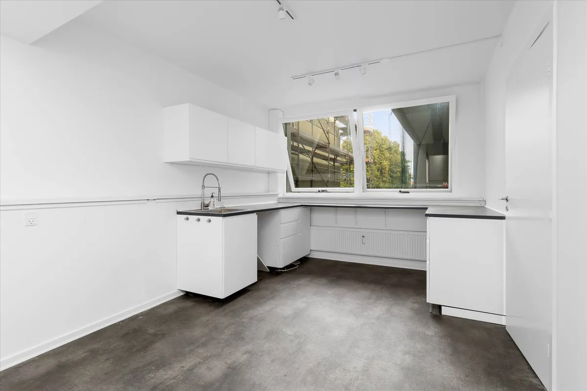 Modernt kontor med hvide vægge, et køkken med en høj skab, en blikfang til vinduet og et hjørne med en radiator