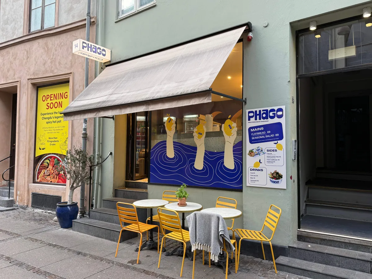 Take-away restaurant med gult stolpebord og terrasse. Lys og luftig, med udendørs bord og tæppe. Lysstofrør og vinduer.