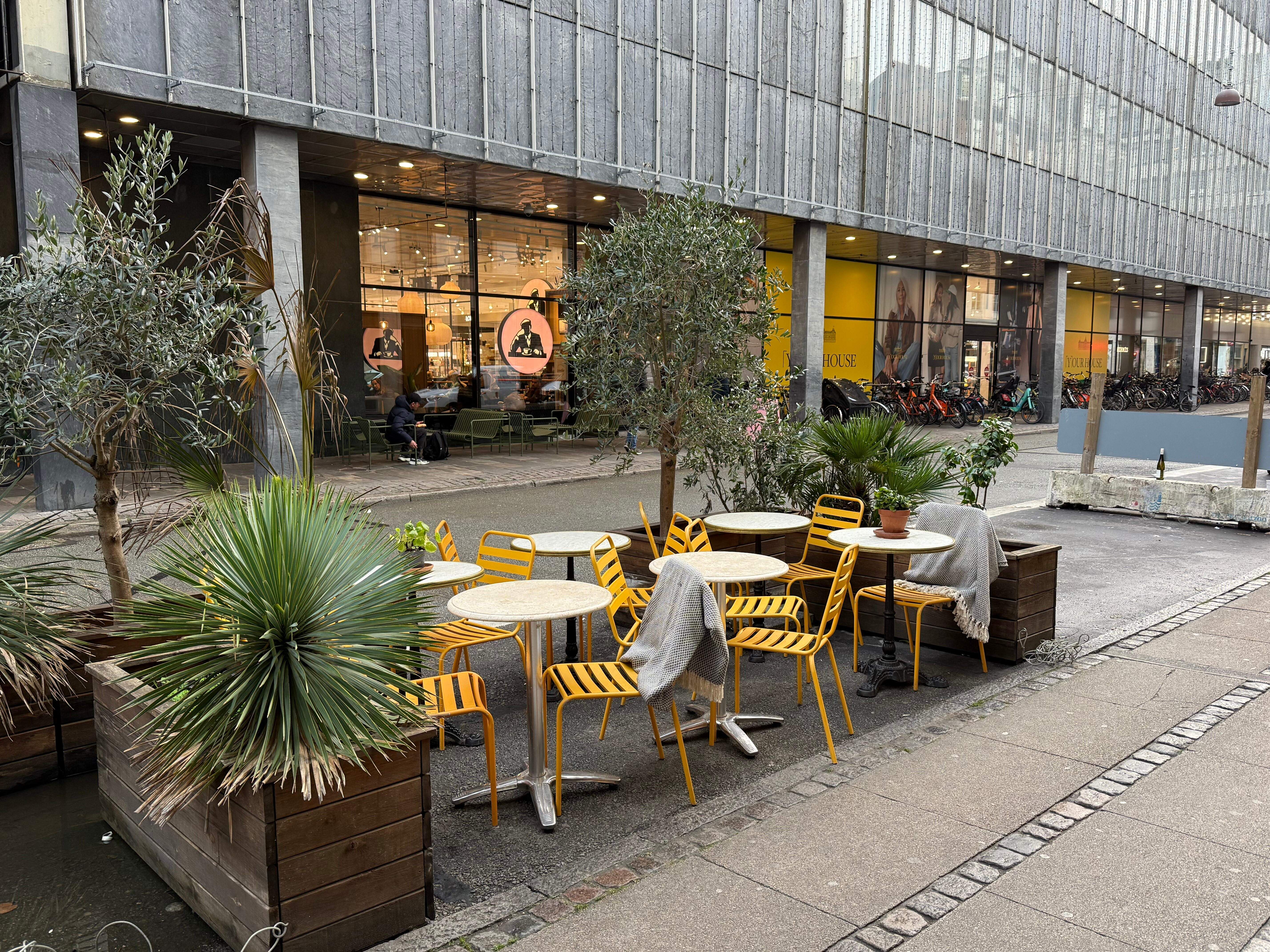 En terrasse med gule stole og borde samt flere plantekasse med grønne planter i forgrunden