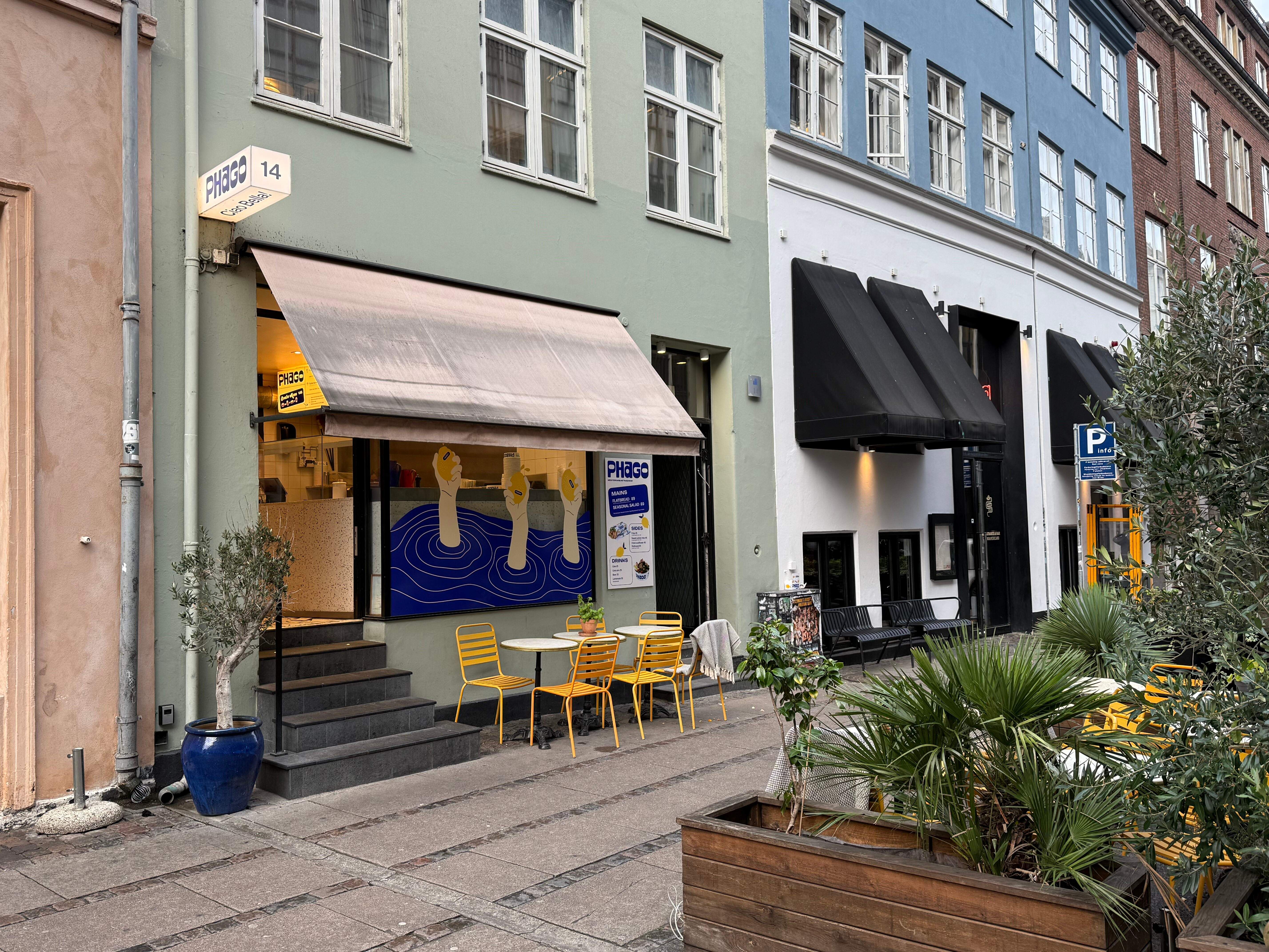 Kaffebar Phago med afkrydsningsfelter, gult bord og stole samt en olivenplante i blå potte