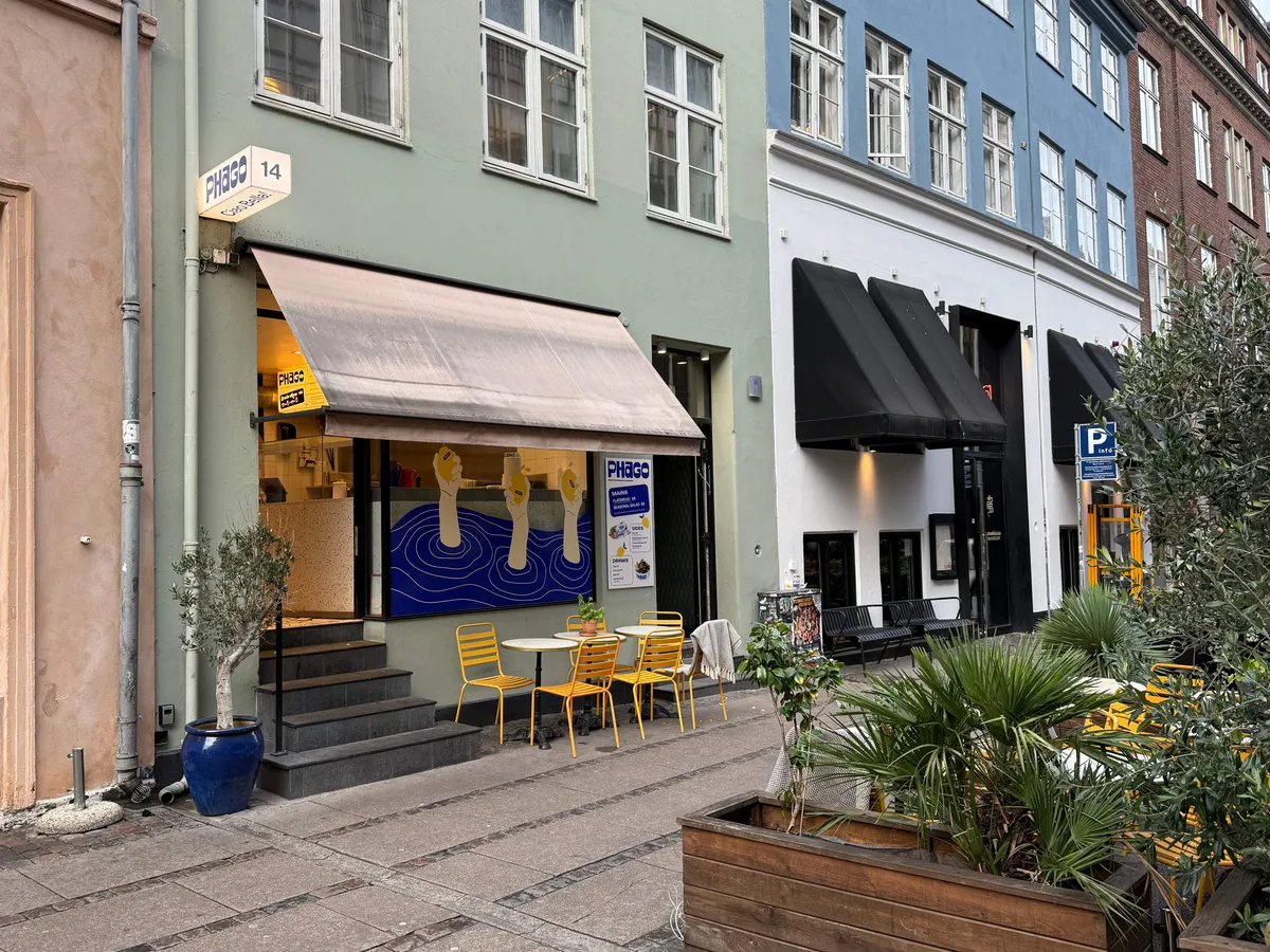 Kaffebar Phago med afkrydsningsfelter, gult bord og stole samt en olivenplante i blå potte