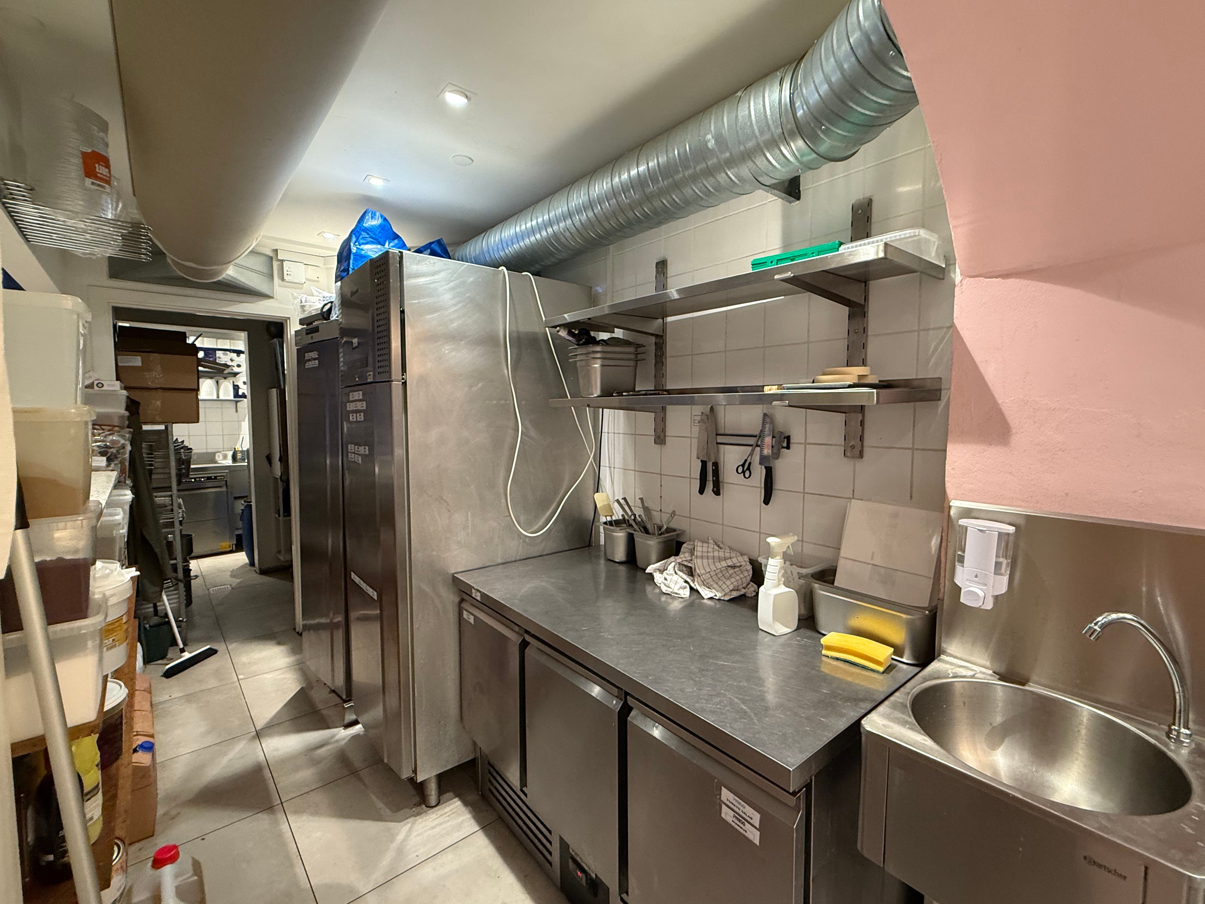 Køkken med flere køleskabe og to stålbænke, hvor der er tøj, skeer, knive og en skraber på den ene bænk.
