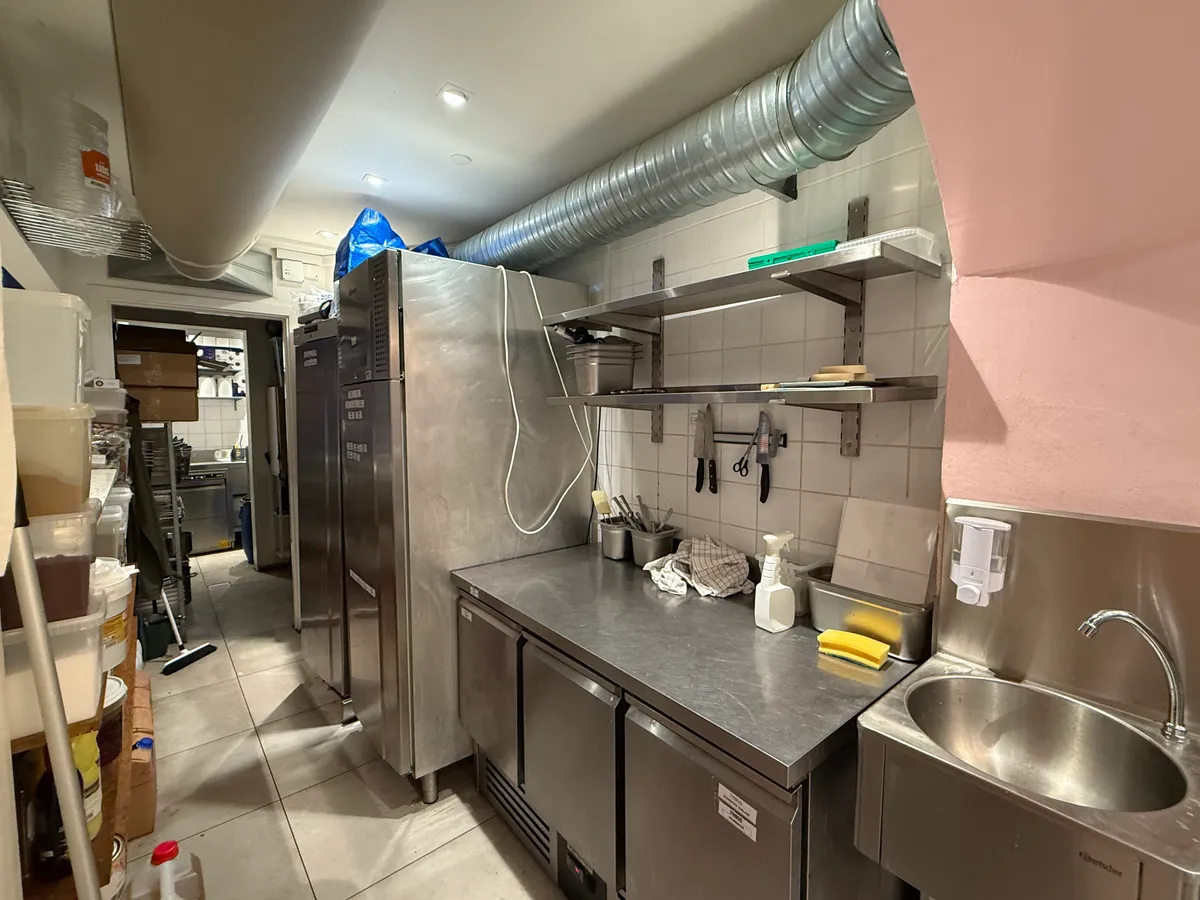 Køkken med flere køleskabe og to stålbænke, hvor der er tøj, skeer, knive og en skraber på den ene bænk.