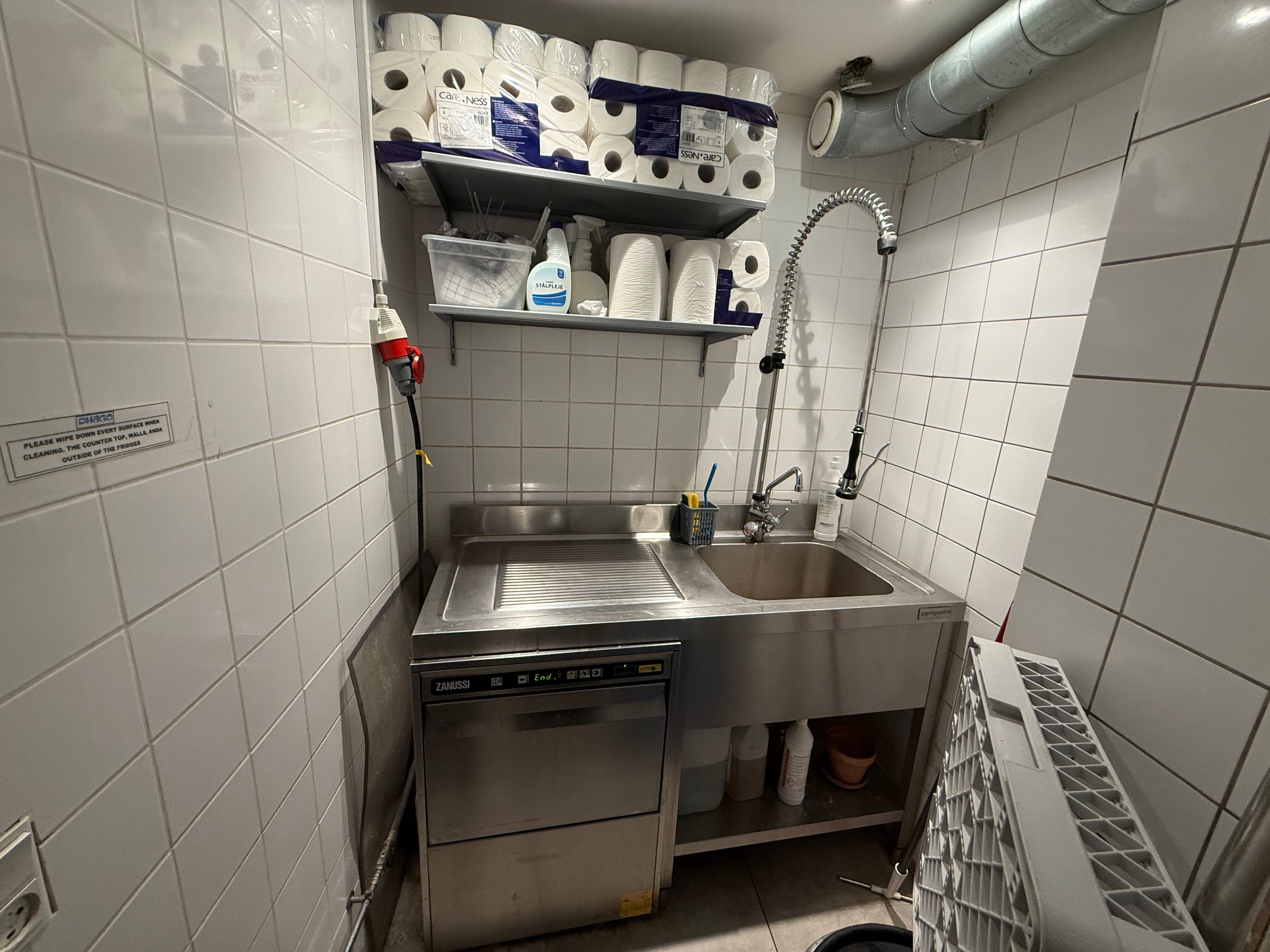 Køkken med to strygebrætter, en maskinvask og en vandhane med to rulleservietter og rengøringsmidler.