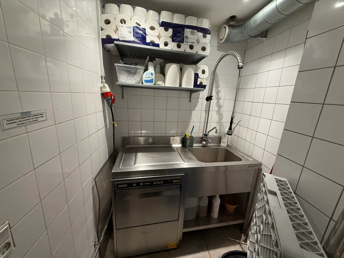 Køkken med to strygebrætter, en maskinvask og en vandhane med to rulleservietter og rengøringsmidler.