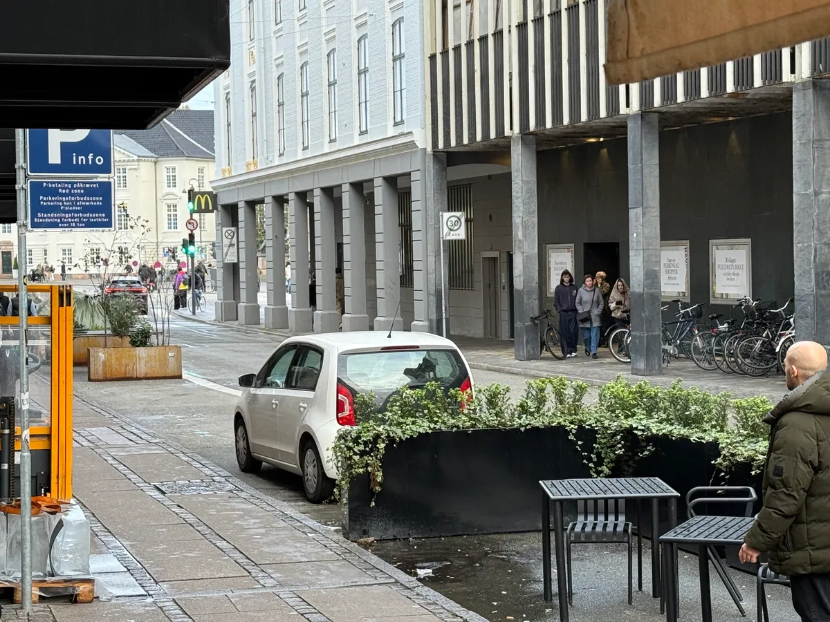 En hvid bil parkeret i en grøn zone med plantager og bord og stole på fortauet