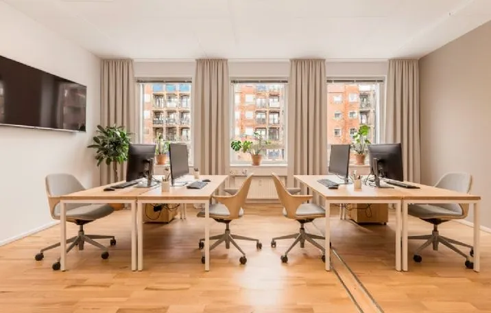 Kontor med trægulve og højloft. To skriveborde med computere og stole. Vinduer med gardiner og bygninger bagved.