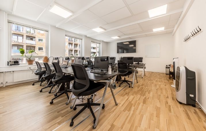 Kontor med lyse lofter og vinduer. Værelse med skriveborde og stole. Luftkøleapparat i hjørnet. Planter på vindueskarmen.