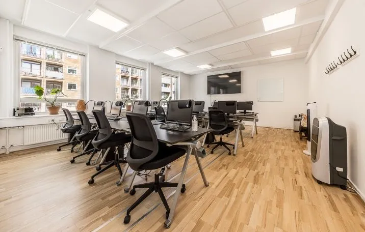 Kontor med lyse lofter og vinduer. Værelse med skriveborde og stole. Luftkøleapparat i hjørnet. Planter på vindueskarmen.