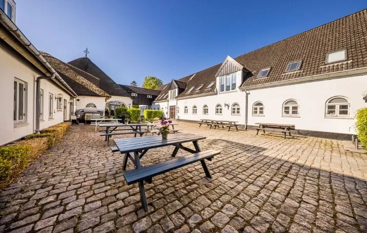 Kontorområde med stolpebord og bænke på stenbrolægget plads i hyggeligt gårdmiljø med røde træhuse og hvide murstensbygninger