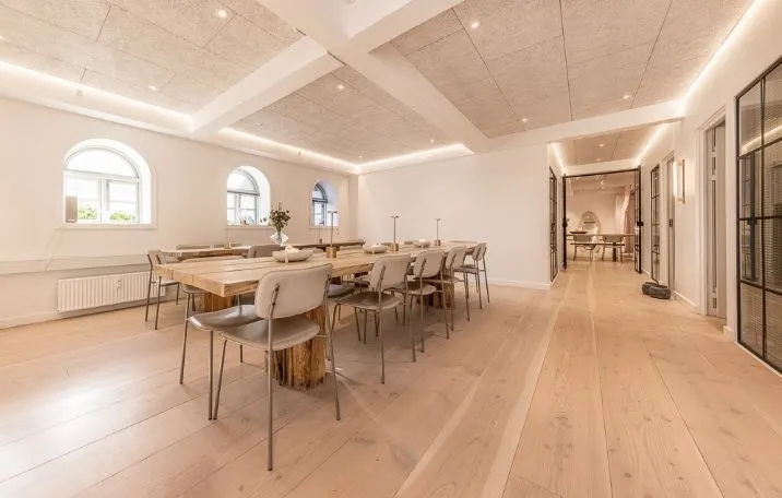 Kontorrum med lyse trægulve og store vinduer, møbler og plante i hvidt og beige