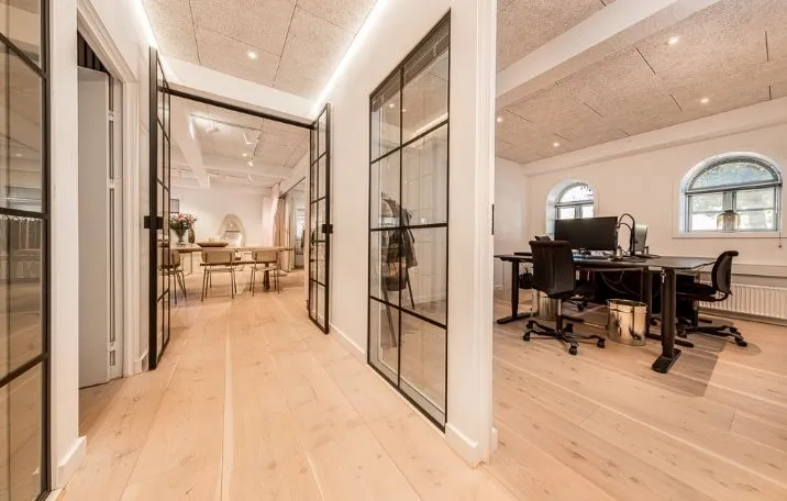 Kontor med lyse gulve, slanke stolpe og glasdøre. Bord med monitor og skrivebordslampe. Højt, halvcirkelformet vindue. Bord