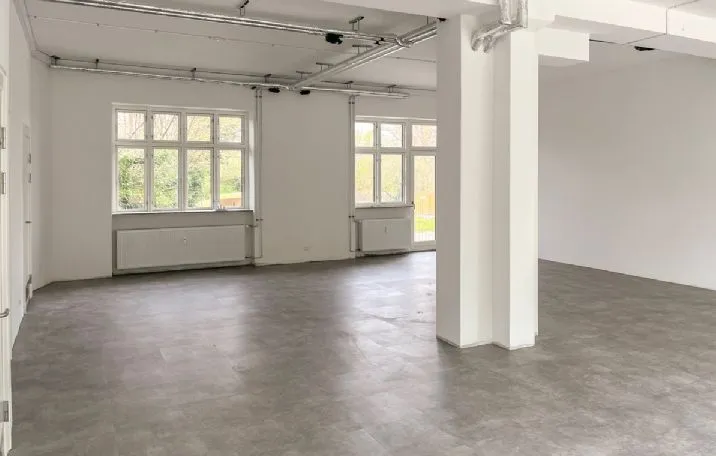 En stor og lys rummeligt lokale med lyse gulvfliser og hvide vægge, hvor der er store vinduer og radiatorer