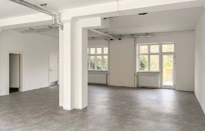 Indenfor en lys og rummelig lejlighed ser man to store vinduer og hvide vægge samt et stort, tømret loft med integrerede