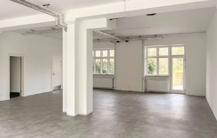 Indenfor en lys og rummelig lejlighed ser man to store vinduer og hvide vægge samt et stort, tømret loft med integrerede