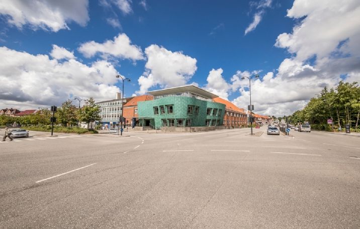 Gadekryds med to biler, flere biler og personer i baggrunden, bygninger med tegltag og facader i grønt og brungul glas og